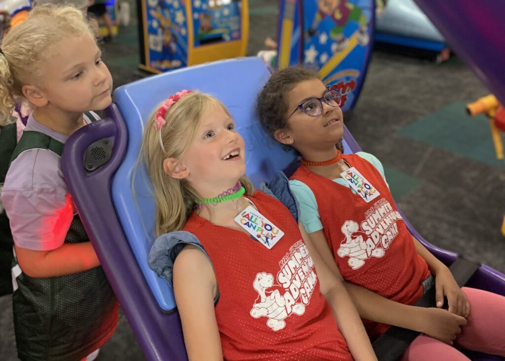 A picture of 3 young girls wearing their Summer Adventure Camp vests and smiling while playing a game at the arcade.
