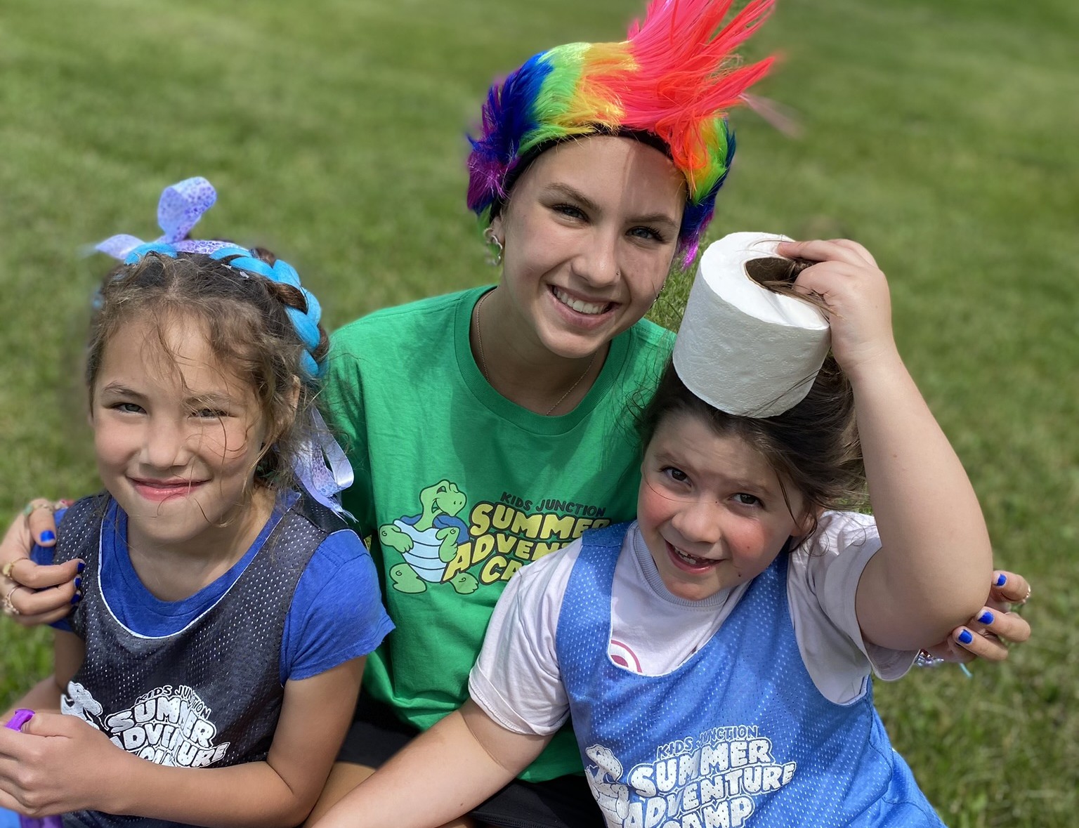 A picture of a Kids Junction Summer Adventure Camp counselor sitting on the grass with two campers wearing their Summer Adventure Camp vests.
