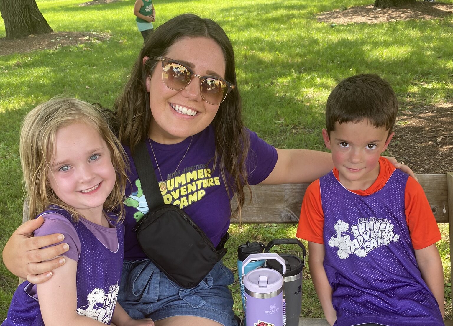 A picture of a Summer Adventure Camp teacher wearing a Summer Adventure Camp t-shirt and sitting on a picnic bench while hugging two campers.