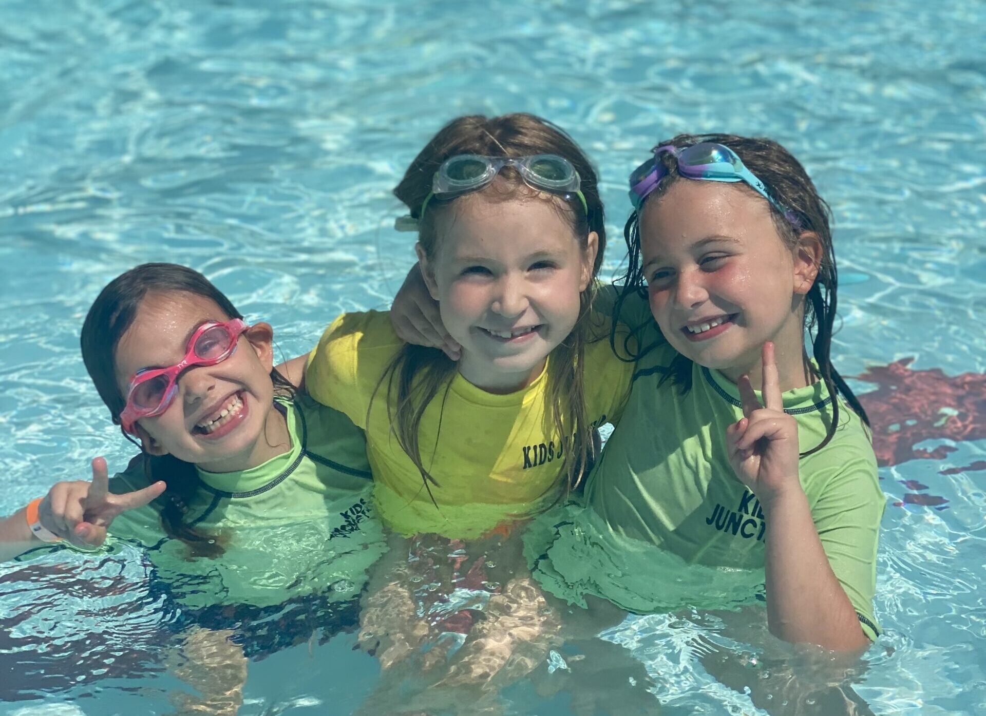 A picture of three little girls hugging while sitting in the pool and smiling at the camera.
