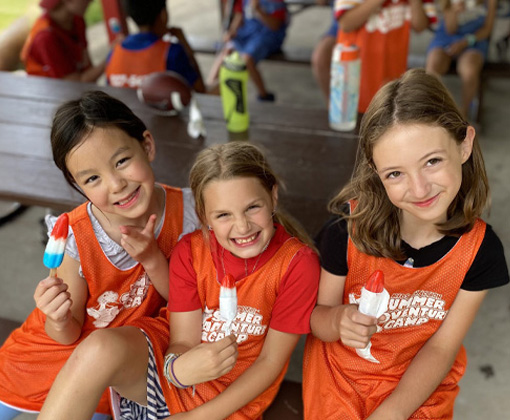 A picture of a Kids Junction Summer Adventure Camp counselor sitting on the grass with two campers wearing their Summer Adventure Camp vests.